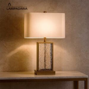 Abajur de vidro com detalhes em metal com cúpula branca sobre uma mesa branca com pés metálicos dourados
