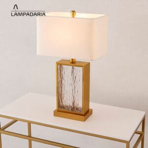 Abajur de vidro com detalhes em metal com cúpula branca sobre uma mesa branca com pés metálicos dourados